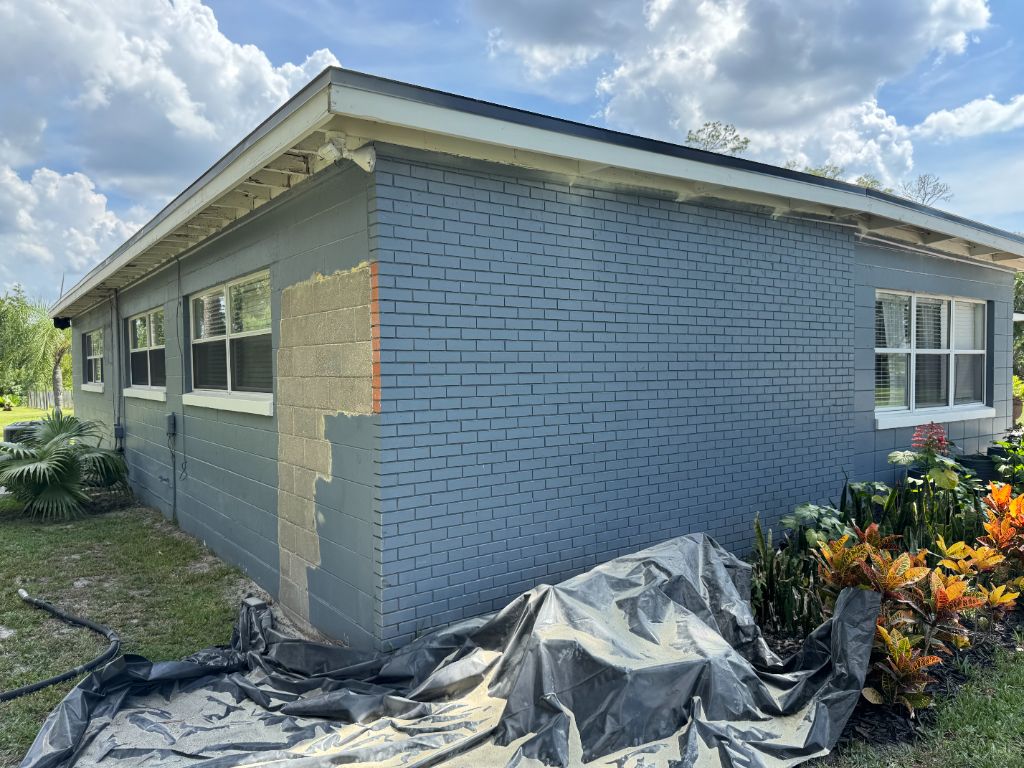 Corner of home during sandblasting — protective sheeting on ground, large areas of red brick already revealed