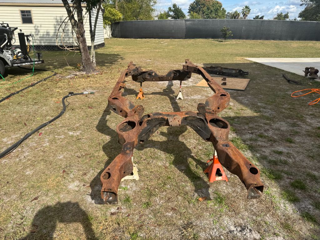 1984 Buick Grand National frame before blasting — heavily rusted full chassis laid out on stands, thick orange-red rust covering all steel surfaces