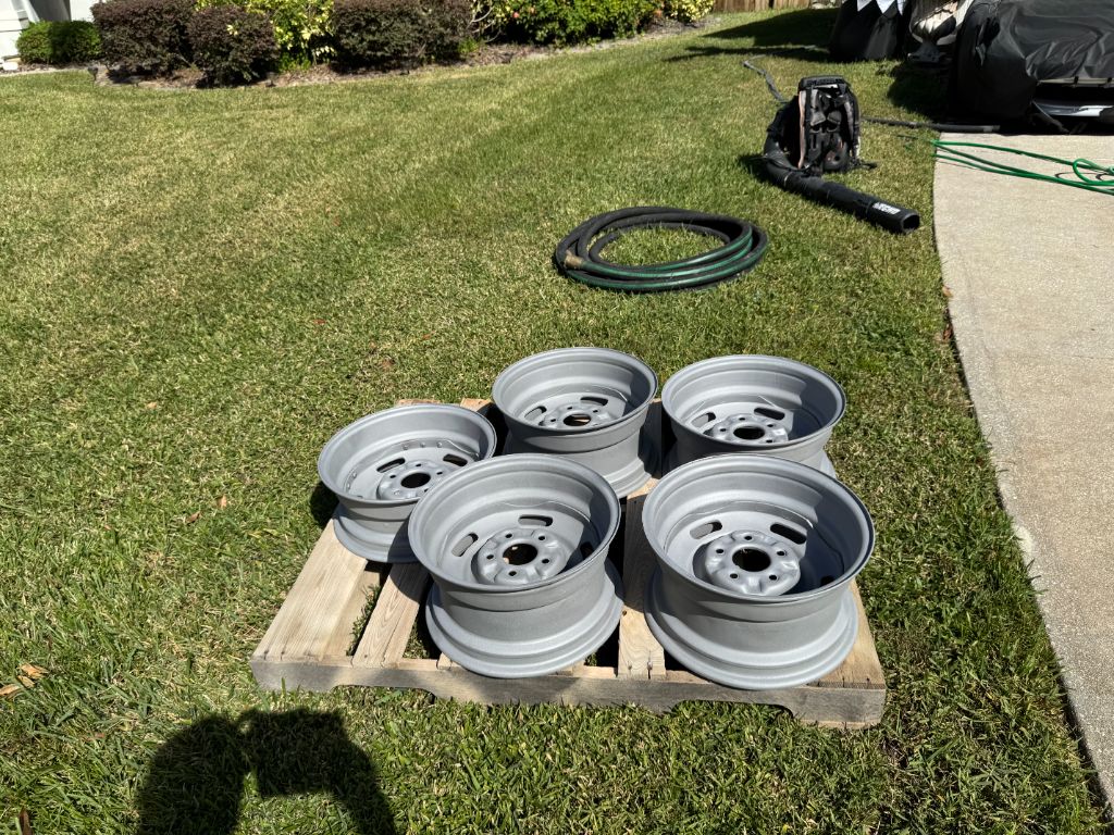 Corvette steel rims after blasting — all 5 wheels clean uniform bare steel gray finish, stacked on wooden pallet, Black Ox blasting rig visible in background