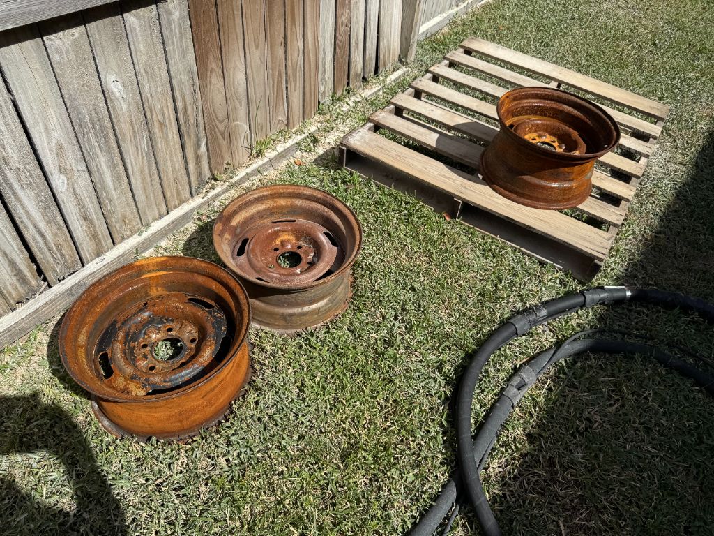 Corvette steel rims before blasting — 3 wheels showing heavy orange-red rust covering entire surface, laying on grass with sandblasting hoses nearby